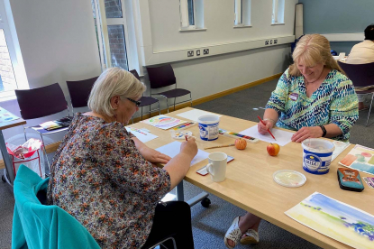 Two women painting together at a table in a bright community room.